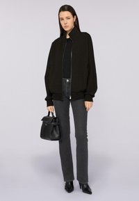 Chaqueta bomber negra con cremallera y cuello alto, llevada sobre una parte superior negra y jeans grises. La modelo sostiene un pequeño bolso negro y lleva zapatos de tacón negros.