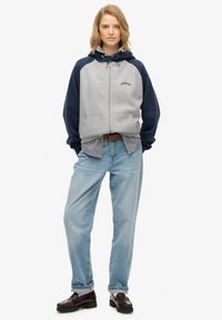 Sudadera gris con cremallera y mangas azul marino, con un logo en el pecho, superpuesta sobre una camiseta de rayas, combinada con jeans azul claro y zapatos oscuros.