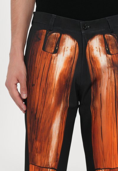 Schwarze Hose mit einem detaillierten orange-braunen Holzmaserungsdruck, Nahaufnahme einer Tasche und einer glatten, matten Oberfläche.