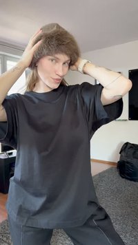 Isik reguleerib pruuni karusnaha mütsi, kandes laia musta särki ja pükse, seismas elutoas, kus on hall vaip ja aken taga.