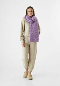 Jonge vrouw die staat, glimlacht, gekleed in beige joggingbroek en sweatshirt, beige instapschoenen en een dikke paarse gebreide sjaal.