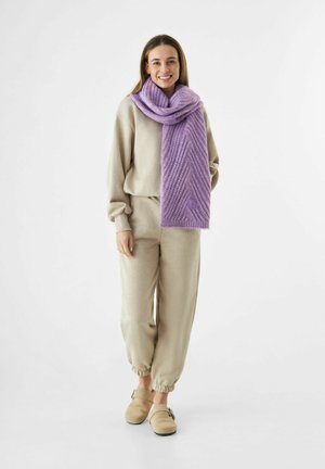 Jeune femme debout, souriante, portant un pantalon de survêtement beige et un sweat-shirt beige, des chaussures à enfiler beiges et une épaisse écharpe en tricot violet.