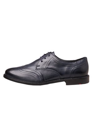 Chaussure habillée pour homme en cuir noir avec détails brogue, fermeture à lacets et talon bas, vue de côté sur fond blanc.