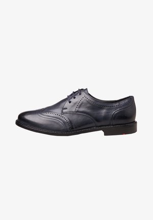 Chaussure habillée pour homme en cuir noir avec détails brogue, fermeture à lacets et talon bas, vue de côté sur fond blanc.