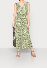Jumpsuit floreale verde con scollatura a V profonda, spalline arricciate, design a gamba larga e cintura coordinata. Motivi beige e verdi su tutta la superficie.