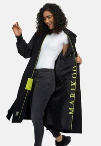 Black long coat with a hood, featuring a yellow zipper and inner lining, textured fabric, and a large yellow logo on the interior.