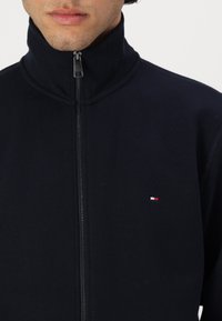 Navy zip-up jacket with a high collar, made of fleece material, featuring an embroidered logo in red, white, and blue on the chest.