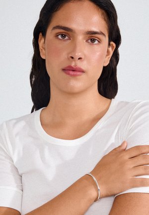 Mladá osoba s tmavými vlasmi, ktorá má na sebe bielu krátkym rukávom košeľu a hladký strieborný náramok, s ramenom preloženým cez hrudník.