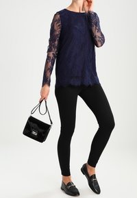 Top in pizzo navy con maniche lunghe trasparenti; abbinato a leggings neri e mocassini neri slip-on. Tiene una piccola borsa nera.