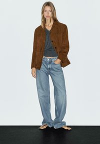 Bruine suède jack, grijs V-hals shirt, losse blauwe denim jeans en zwarte slippers, staand op een donkere ondergrond tegen een lichte achtergrond.