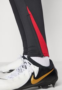 Sorte atletiske tights med en rød side stripe, parret med hvide og sorte fodboldstøvler med et gul Nike-swoosh. Tekstureret stof synligt.