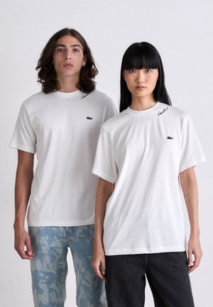 Due persone stanno in piedi di fronte, indossando t-shirt bianche Lacoste, jeans a camuflaggio blu chiaro e jeans neri, su uno sfondo bianco uniforme.