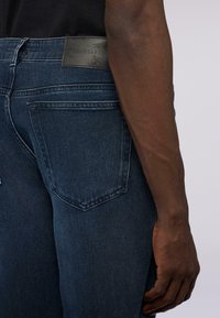 Jeans de mezclilla azul oscuro con una etiqueta de cuero negro etiquetada "Trussardi". Las características incluyen costuras sutiles y un diseño clásico de cinco bolsillos.
