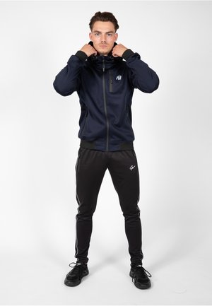 Marineblauwe zip-up jack met een aansluitend ontwerp, inclusief een capuchon en ritszakken. Zwarte tapered broek met witte zijstrepen en logo-accent.
