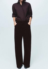 Camisa burdeos de botones con mangas remangadas, combinada con pantalones de pana burdeos de pierna ancha con bolsillos frontales, usados con zapatos negros.
