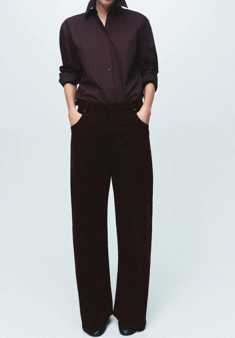 Camisa burdeos de botones con mangas remangadas, combinada con pantalones de pana burdeos de pierna ancha con bolsillos frontales, usados con zapatos negros.