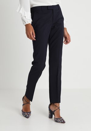 Pantalon de tailleur bleu marine à coupe slim, avec une taille plate et des passants de ceinture. Associé à des chaussures à talons hauts à imprimé floral et un chemisier blanc à volants.
