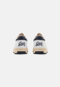Weiße Sneakers mit cremefarbenen Akzenten, strukturiertem marineblauen Absatz und gesticktem Markennamen auf der Rückseite. Gummisohle mit leichtem Profil.