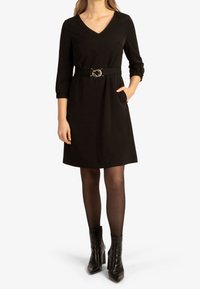 Schwarzes V-Ausschnitt-Kleid mit Dreiviertelärmeln, tailliert mit einem metallischen Gürtel und Seitentaschen. Kombiniert mit schwarzen Stiefeletten.