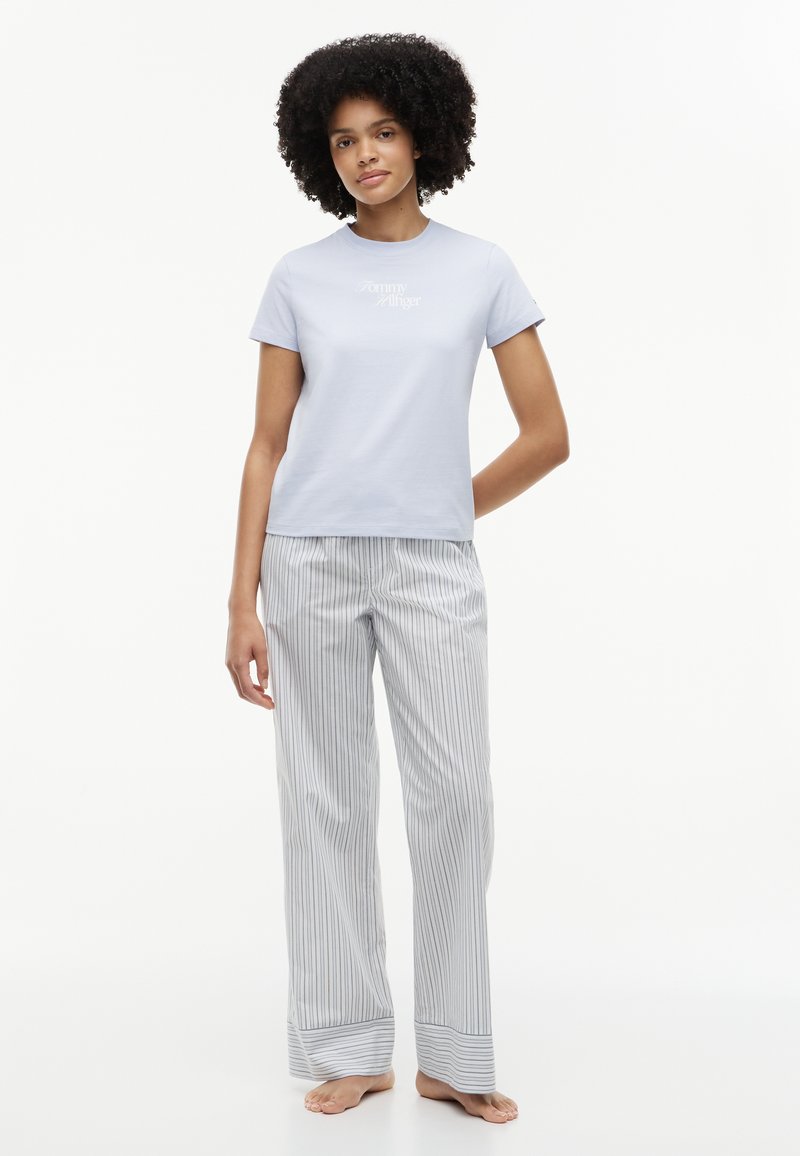 Femme debout pieds nus portant une chemise bleu clair à manches courtes et un pantalon de pyjama blanc avec de fines rayures verticales grises.