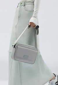 Grå crossbody-väska med rektangulär form, slät textur och en framficka. Utrustad med en avtagbar rem med justerbar hårdvara.