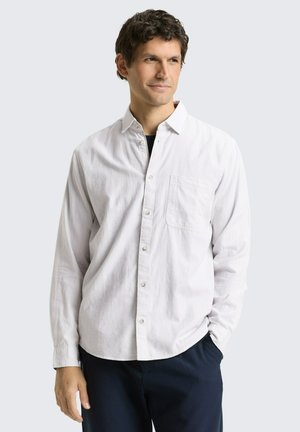 Mężczyzna z krótkimi kręconymi włosami, ubrany w białą koszulę zapinaną na guziki z kieszenią na piersi oraz ciemne spodnie, stojący z jedną ręką w kieszeni.