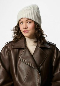 Berretto a coste crema, texture morbida, abbinato a una giacca di pelle marrone scuro con colletto a scollatura e dettaglio con zip.