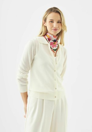 Vrouw in een witte cardigan en wijde broek draagt een kleurrijke bloemige sjaal om de hals tegen een witte achtergrond.