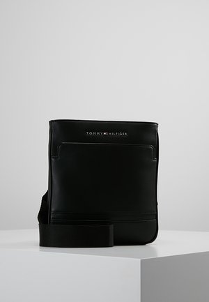 Schwarze Leder-Tommy Hilfiger Crossbody-Tasche mit Fronttasche und verstellbarem Stoffriemen auf einer weißen Oberfläche vor einfachem Hintergrund.