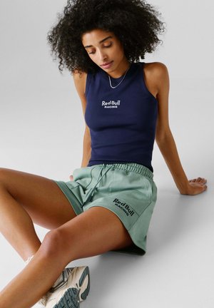 Marineblaues ärmelloses Oberteil mit dem Schriftzug "Red Bull Racing", kombiniert mit mintgrünen Shorts, die dasselbe Logo tragen. Leichte Sportsneaker vervollständigen das Outfit.