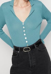 Cardigan turchese a coste con scollatura a V e sei bottoni bianchi, abbinato a jeans neri a vita alta e una cintura nera con una fibbia d'argento.