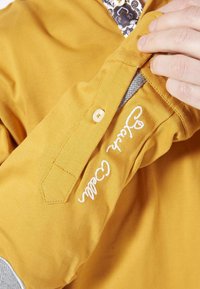 Chemise jaune à col boutonné, avec des accents gris et un logo brodé blanc sur la manche, texture lisse.