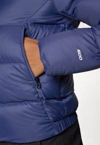 Námornícka modrá páperová bunda s prešívaným vzorom, obsahuje zipsovú kapsu a elastické manžety. Ľahký materiál s lesklým povrchom.