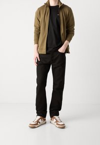 Man i olivgrön zip-jacka, svart t-shirt, svarta byxor och vita sneakers med bruna detaljer som står mot en enfärgad bakgrund.