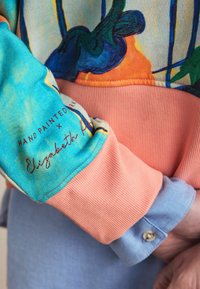 Kleurige trui met een blauw, oranje en aqua ontwerp en geribbelde roze manchetten. De stof toont een handgeschilderd patroon en een geborduurde naam.