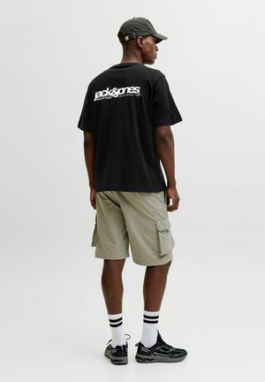 Homme portant une casquette noire, un t-shirt noir avec un logo blanc dans le dos, un short cargo beige, des chaussettes blanches à rayures noires et des baskets noires, debout de profil.