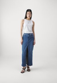 Hvit ermeløs topp med logo, høy midje blå denim skjørt med splitt foran, og svarte blokkhæl sandaler. Jevn tekstur generelt.