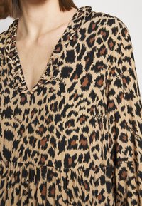 Femme portant une robe beige avec un motif léopard marron et noir et un col en V à volants, montrant le haut du torse et le décolleté.