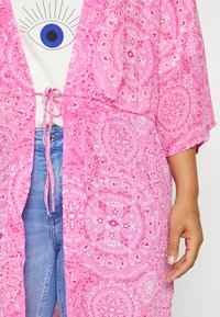 En närbild av en person som bär ett rosa mönstrat plagg över en vit skjorta med ett ögonmönster, tillsammans med blå jeans.