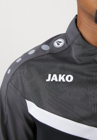 Grauer Sportjackett mit einem strukturierten Muster, weißen Akzenten und Logo. Ausgestattet mit kreisförmigen Verzierungen an der Schulter.