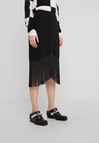 Černá sukně s třásněmi na lemu, vyrobená z hladké látky. V kombinaci s černými dřeváky se metalickým akcentem na popruhu.
