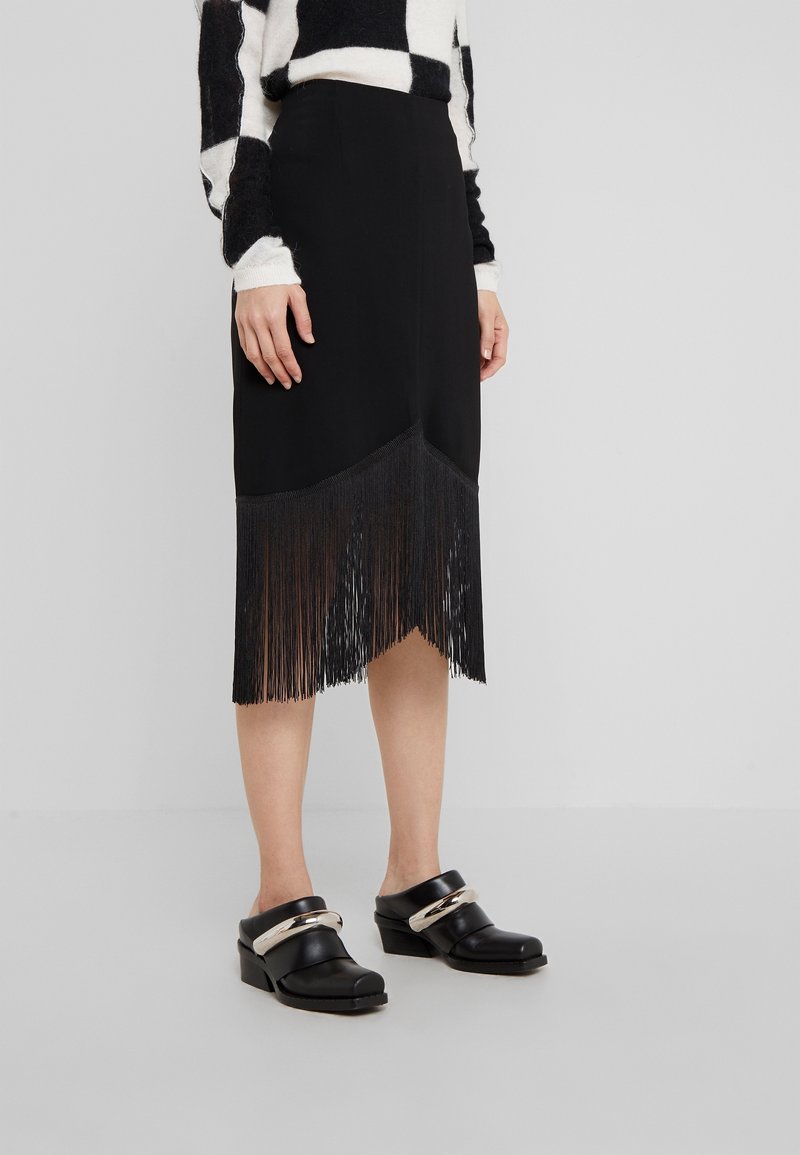 Černá sukně s třásněmi na lemu, vyrobená z hladké látky. V kombinaci s černými dřeváky se metalickým akcentem na popruhu.