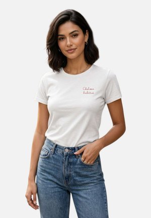 Donna con capelli scuri fino alle spalle indossa una maglietta bianca a maniche corte con testo rosso e jeans blu a vita alta, posando con una mano in tasca.