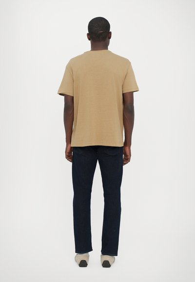 T-shirt à manches courtes beige en coton, associé à un jean en denim noir. Le jean a une coupe droite et une légère texture, avec des chaussures décontractées.