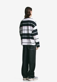 Mand står med ryggen til iført en langærmet skjorte med sort og hvid vandret stribe, løse sorte jeans og grå sneakers på hvid baggrund.