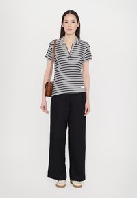 Mustavalgeline triibuline polo särk, millel on sälguga krae, koos lahtiste mustade pükstega ja pruuni õlalokiga kotiga. Naine seisab neutraalse tausta ees.