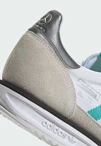 Detailný záber na pätu tenisky Adidas SL '72 s béžovým semišom, bielou podrážkou, tyrkysovým pruhom a strieborným logom Mercedes-Benz AMG na golieri.