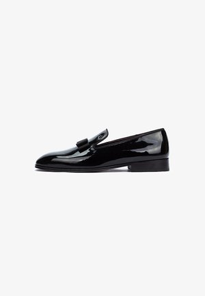 Zapato negro de charol de tipo slip-on con tacón plano, que presenta un adorno decorativo en forma de lazo y un diseño elegante y minimalista.