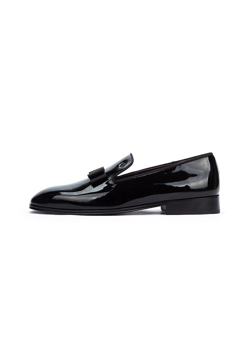Zapato negro de charol de tipo slip-on con tacón plano, que presenta un adorno decorativo en forma de lazo y un diseño elegante y minimalista.