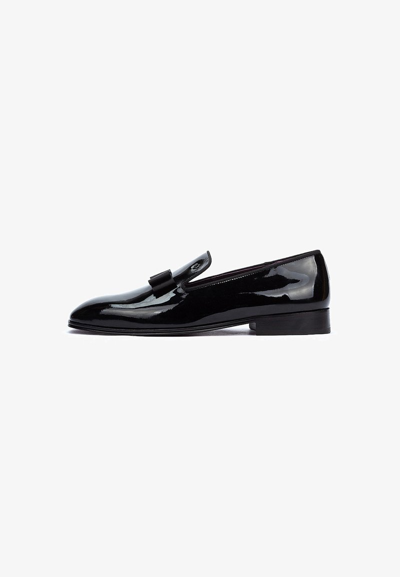 Zapato negro de charol de tipo slip-on con tacón plano, que presenta un adorno decorativo en forma de lazo y un diseño elegante y minimalista.